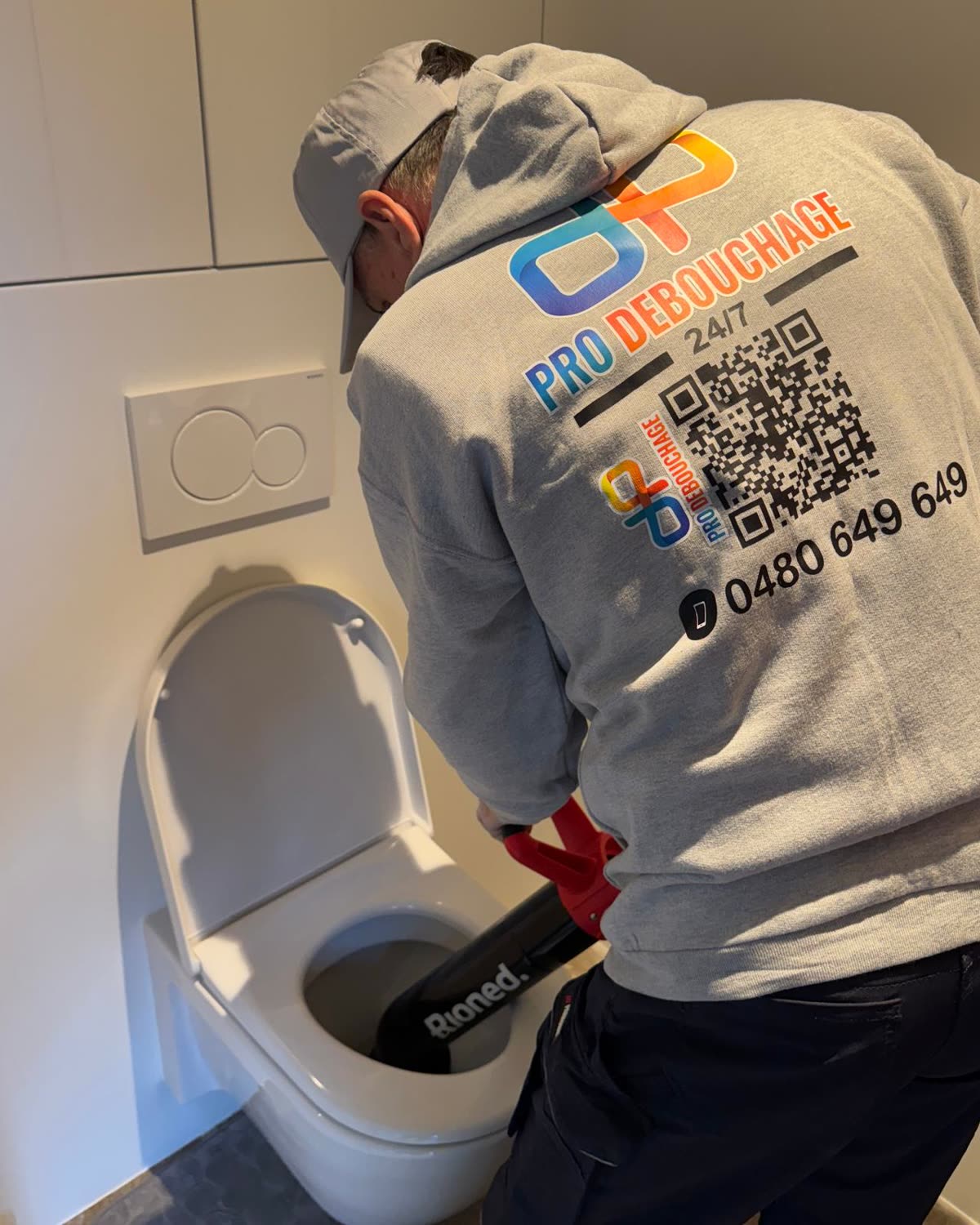Technician unblocking a toilet with a Rioned drain rod, Pro Débouchage jacket.